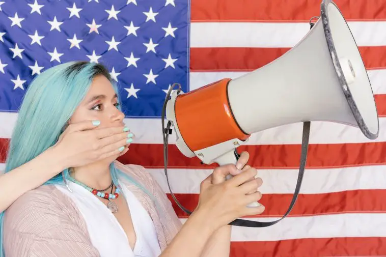Woman holding megaphone with hand over mouth in front of American flag.